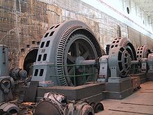 Interior of building showing three old fashioned vertical generators, like an upright wheel, each more than twice the height of a person.