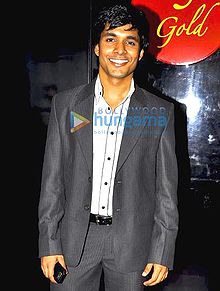 An Indian man smiling, wearing a dark grey suit, with a white shirt under and holding a mobile in his right hand.