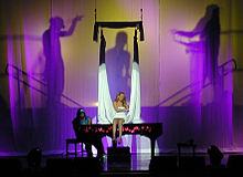 A blond woman sits atop a piano and sings. A long, white cloth hangs around her while a silhouette behind the woman shows two male figures as if holding the white cloth.