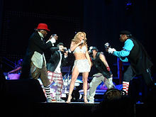 A blond woman singing, wearing a white top and short skirt. She is flanked by four boys, who pretends to be taking her picture with camera.