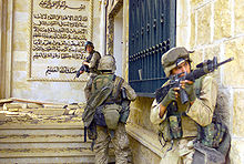 photograph of three Marines entering a partially destroyed stone palace with a mural of Arabic script