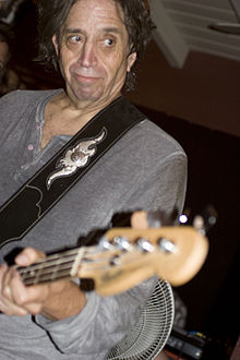 Mark Tulin—a middle-aged Caucasian man with long brown hair in a grey shirt–plays a bass guitar.