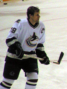 An ice hockey player skating on the ice. He wears a white jersey with black trim and is helmetless. He looks forward and holds his stick horizontally across his torso in a relaxed fashion.