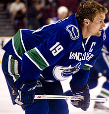 A Caucasian ice hockey player in his mid-thirties. He stands crouched over on the ice with his stick rested horizontally on his knees. He wears a blue jersey with white and green trim and is helmetless.