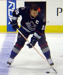 An ice hockey player stickhandling the puck. His head is down and his skates are shoulder-width apart. Wearing a black jersey, he is helmetless.