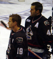 Two ice hockey players are standing next to one another and looking forward. Neither are wearing their helmets. They wear black jerseys.