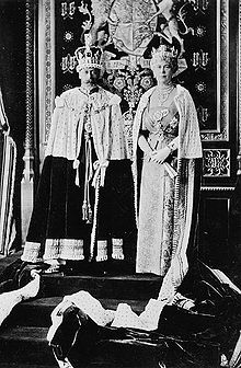 Late-middle-aged couple in crowns and ermine capes stand on a dais