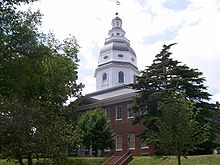 A colonial-style brick building with a colonnade and a large grey and white dome topped by a flagpole with the Maryland and United States flags sits behind a grassy lawn, shielded by mature oak trees