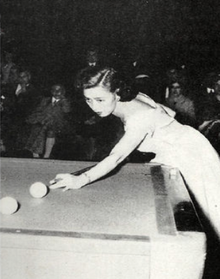 Black-and-white photo of Japanese woman in fancy dress, approximately 30 years old, stretched out over corner of carom billiards table with her cue stick in hand. Her eyes are intently focused on the shot in front of here, two billiards balls about a foot apart, with her cue tip about two inches behind the closest ball in position for the hit; the table scene is in spotlight and in the much darker background can be just made out spectators in chairs, all appearing to be men.