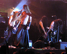 Four long haired musicians perform on a small stage in the spotlight, long hair swinging. The singer, wearing a black leather floor-length skirt,  and the bass guitar player are bare chested with black leather wrist bands. The two other guitar players are in sleeveless black shirts, one with knee-length jeans and the other with black wrist band. Amplifiers crowd the stage.
