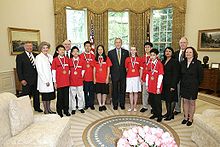 Fourteen people stand in a line, seven of whom are wearing medals and red t-shirts and the other seven of whom are dressed formally.