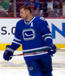 An ice hockey player, visible from the knees upwards, skating with spectators visible behind him. He is holding a hockey stick and wearing gloves and a sweater with a simple drawing of a hockey stick on the front, and a large "A" below his left shoulder. Not wearing a helmet, he is looking to his right, and has a relaxed expression.