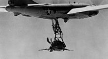 Black-and-white photograph shows diminutive jet fighter suspended from a large aircraft in flight, through a trapeze.