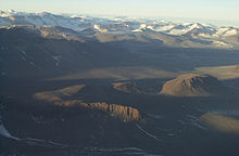  In the foreground is a landscape of dull brown mounds and undulations, behind which are snow-covered mountain peaks.