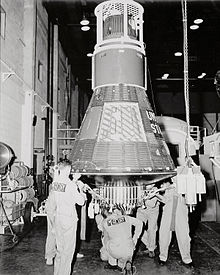 Men in overalls clustered around the base of an upright conical spacecraft, which is two or three times taller than them, inside a large room