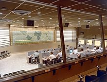 A large room seen through windows at the rear, with two rows of people seated at desks and computers, and a map taking up the whole of the front wall.