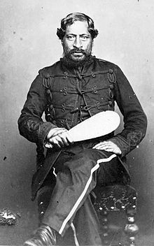 Formal seated portrait photograph of a man in his 50s wearing a uniform and holding a patu.