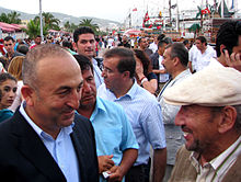 Two older Turkish men stand facing each other, one bald, the other wearing a white cap, while a large crowd mingles behind them along a waterfront.