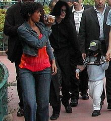 A group of adults and a child are shown in the photo. An African American female with short brown hair who is wearing a jean jacket and a light wash pair of jeans with a red shirt is seen holding a video camera, which is pointing forward. To the right of the female there is an opened black umbrella that is held above a light-skinned male with long black hair who is wearing all black clothes. In front of the male there is a child with black hair that is wearing a black cap with a blue shirt, a pair of white pants and black shoes. To the farthest right there are two Caucasian males with dark brown hair. In the background, trees, bushes and people can be seen.
