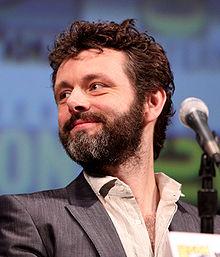 A bearded man wearing a suit is pictured smiling behind a microphone.