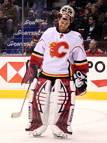 a hockey goaltender in a white jersey with a stylized red C logo on his chest and his mask raised above his forehead stares down at the ice as he is skating.