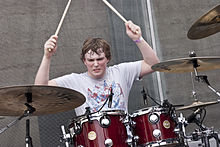 Mike Byrne—a Caucasian male 21 years old with short brunette hair with a grey t-shirt—drums behind a red drum kit.