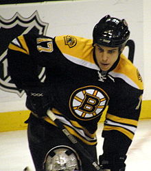 A frontal view of a hockey player in his early twenties, cut off at the thighs, during a hockey game.  He is wearing a black and yellow jersey with a logo containing a capitalized "B". The number "17" is visible on his jersey's left sleeve.  He is looking downwards with an intent expression on his face.