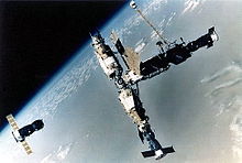 An image of a space station consisting of four modules arranged in a T-shape. A short, stubby module is docked to a longer, stepped-cylindrical module which has a number of docking ports arranged in a sphere at one end. Two other modules, similar in size, project from opposing ports on this sphere. A Progress spacecraft is docked to the short module, a Soyuz spacecraft to the end of the lower module in the crossbar of the T, and another Progress spacecraft is seen a distance away from the module cluster, carrying out undocking operations.