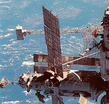 A partial view of a space station, with two modules visible. The vertically aligned module has a large, dish-shaped antenna projecting from it, and a large truss can be seen horizontally behind it, with a white box-shaped device mounted to the end. The horizontal module has a smaller truss mounted to the end of it, with a white backpack attached to this. The module also features two large solar arrays and a number of cameras mounted to a unit on the distal end of it. Another solar array is visible behind this module.