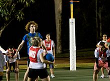 Woman holding a netball while other players stand watching her. Woman and shooting team is wearing white with red bibs. Opposing team is wearing blue with green bibs.