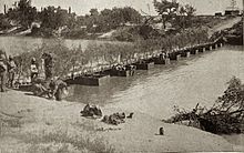A low bridge over twelve pontoons, with troops on the nearby bank