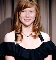 A sixteen year old redheaded caucasoid female is wearing a black halter top, sitting in front of a neutral-coloured cloth background, looking off to the photographer's left.