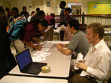 Two young white men sit at a table with several teenage African American students mingle around it, and one signs a paper on it. Also on the table is a laptop.
