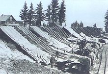 Men working next to chords of wood at the bottom of wooden ramps on the side of a hill. A building and set of trees is on top of the hill. A rail line parallels the chords of wood.