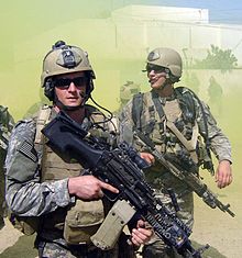 Michael Monsoor, a white male and another white male team-mate, dressed in green camouflage uniform loaded with green combat uniforms. Both are carrying weapons and wearing sunglasses. There is a white building and green smoke in the background
