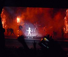Faraway image of a stage lit by red light and backdrops of flames. A potlight falls on a female blond woman in the centre who wears a large white headdress and holds a stick in her left hand. She is surrounded by similar dressed male dancers who also wear a half-sleeved jacket.