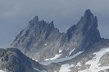 Lightly glaciated mountainous landscape with a steep vertical rocky mountain towering above the surrounding terrain in the background.