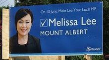 A billboard with a blue background. In white font, up the top of the board is written 'On 13 June, Make Lee Your Local MP' below which is a ticked box, and the words 'Melissa Lee'. Beneath that 'Mount Albert' is written. In the bottom-right corner is the word 'National' with red stars in the southern cross formation at the top-right of the 'N'. To the very left of the billboard is a Korean woman wearing a black shirt.