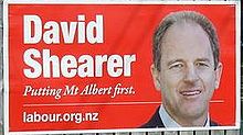 A billboard with a red background, and white font reading 'David Shearer'. Below that reads 'Putting Mt Albert first'. At the bottom 'labour.org.nz' is written. To the right is a man with balding brown hair, smiling, and in a white shirt with a black jacket