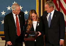 A color image of Murphy's parents standing next to President George Bush in front of an American flag. They are holding Murphy's Medal of Honor in a display case and are looking down at it.