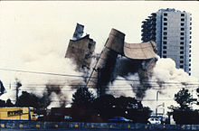The Alfred P. Murrah building is being demolished, and the image shows the building in mid-collapse. A Ryder truck is visible at the bottom left, and the Regency Towers building can be seen in the background at the far right. The demolition has created large clouds of dust that take up a portion of the image.