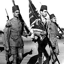 Three men in military uniform moving to the right. The middle one is holding a flag, while the others are holding rifles