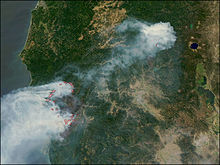 An aerial image of southern Oregon and northern California. The Pacific Ocean is to the west, while the circular Crater Lake and Upper Klamath Lake are visible to the east. Fires are outlined in red. The Biscuit Fire—located in the southwestern corner—forms a large irregular ring shape, and a massive gray smoke plume rises from it. The smoke is drifting southwest over the ocean.Another smaller fire is located to the northeast.