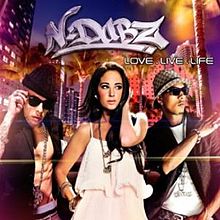 A portrait of three young adults walking in an urban environment at night. On the left and right are young men wearing sunglasses, and in between them is a brunette in a white dress. Above them is a N-Dubz logo in white and below it is the title 'Love.Live.Life' in white capital letters.