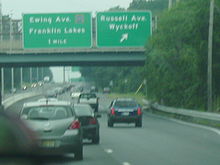 A congested four lane highway with a center concrete barrier containing lampposts that is surrounded by trees. Two overhead signs stand over the roadway with the left sign reading Ewing Ave. County Route 502 Franklin Lakes 1 mile and the sign on the right reading Russell Avenue Wyckoff arrow pointing to the upper right. A highway overpass lies behind the sign display.