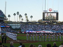 Two rows of men stand on a baseball field holding baseball caps over their hearts. A row of men in gray baseball uniforms and red caps are to the right of the image, while men in white baseball uniforms and blue caps are to the left. The stands are full with crowd members, and other people are standing in the outfield, seen in the background.