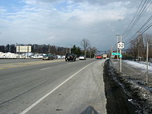 A four-lane highway with a striped median is bordered on both sides by snow-covered areas. A NY 32 shield and reference marker are mounted on a pole to the right of the road.