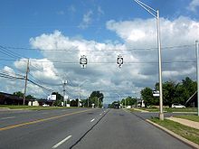 A four-lane highway proceeds into the background, where it disappears from view after descending a hill. Buildings home to various businesses, streetlights, and sidewalks line both sides of the highway. Two overhead signs held up by wires instruct drivers destined for NY 22B southbound to continue straight and those destined for NY 3 westbound to turn right ahead.