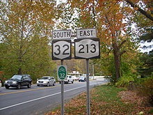A two-lane highway in a forested area during autumn. In the foreground is a sign assembly reading "south NY 32 east NY 213".
