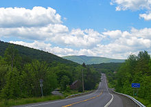 A two-lane highway descends into a valley containing a thick forest. Large ridges are visible to the near far left and in the background; in the distance is a peak rising above of the ridge.